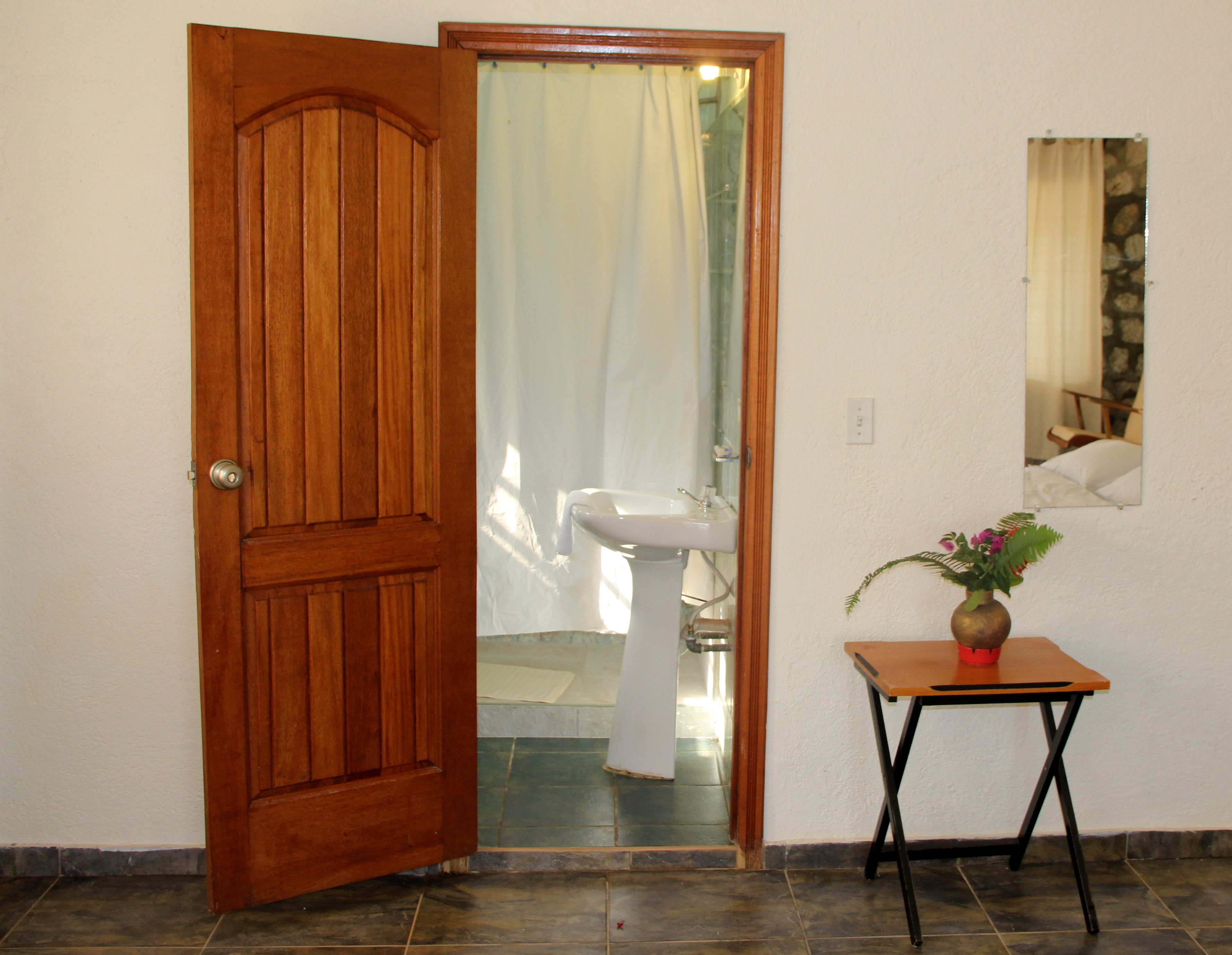 Una puerta de madera que da a un baño con un lavabo blanco y una pequeña mesa con un jarrón.