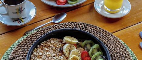 A breakfast table with a bowl of cereal topped with fruits, a plate of sliced fruits, a cup of coffee, a jug of milk, a glass of orange juice, and a basket of bread.