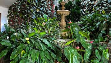 Una fuente de piedra rodeada de plantas y enredaderas exuberantes, frente a un muro con diseño ornamental.
