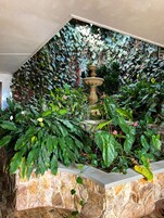 Una fuente de piedra rodeada de plantas y enredaderas exuberantes, frente a un muro con diseño ornamental.
