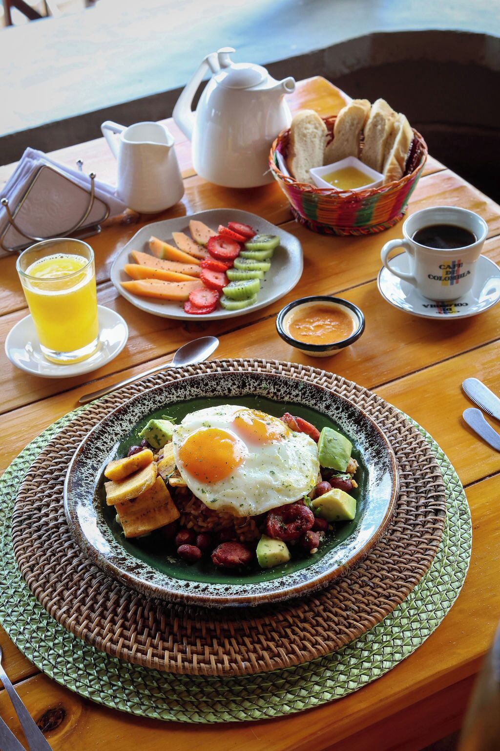 A breakfast setting with a plate of eggs, avocado, and beans, a cup of coffee, and a basket of bread.