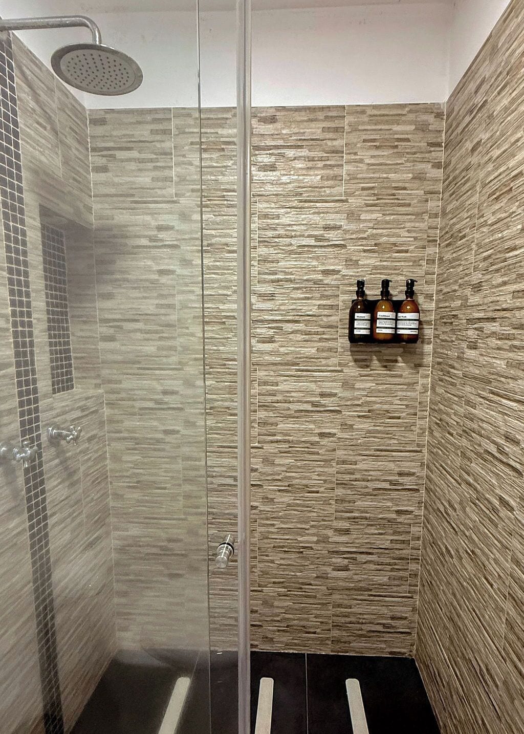 Une douche avec une porte en verre, un pommeau de douche à main et un porte-bouteilles fixé au mur, pouvant accueillir trois bouteilles.