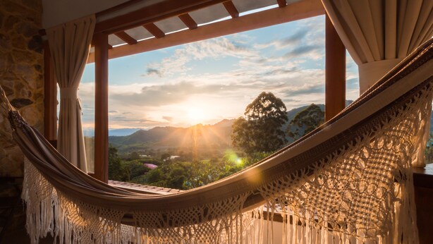 A hammock with macrame fringe hanging in a room with a view of a landscape and mountains. - Hotel Spa La Colina (Pereira)