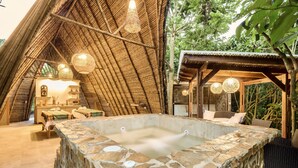 A stone hot tub in a wooden structure with a thatched roof, surrounded by wicker furniture and a small wooden building. - Hotel Spa La Colina (Pereira)