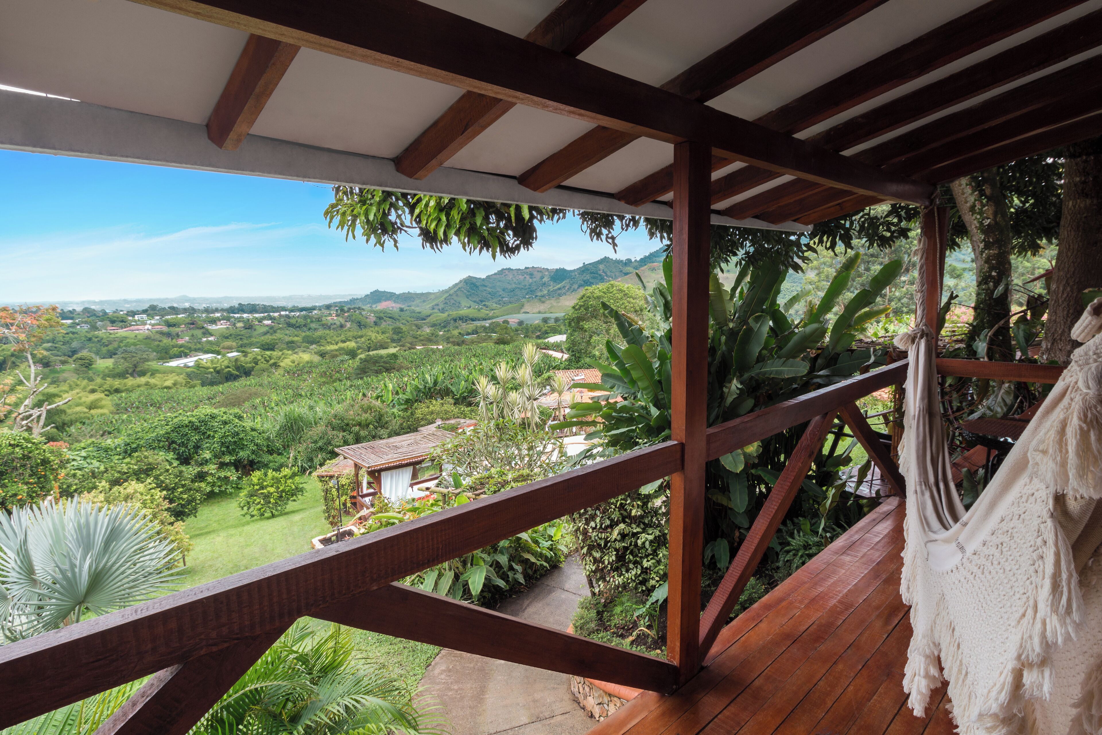 Una terraza de madera con vistas a un paisaje exuberante, que incluye una casa y montañas a lo lejos.