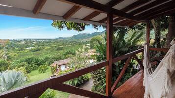 Una terraza de madera con vistas a un paisaje exuberante, que incluye una casa y montañas a lo lejos.