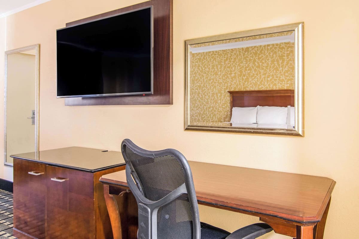 A hotel room with a wooden desk, a chair, a flat-screen TV mounted on the wall, and a framed mirror reflecting a bed with white linens.