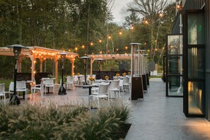 Un patio al aire libre con sillas y mesas blancas, luces colgantes y una estructura de pérgola.