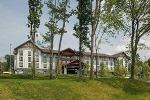 Un hotel con diseño arquitectónico moderno, que cuenta con una entrada central con pasillo cubierto, varios pisos y un estacionamiento visible en primer plano.