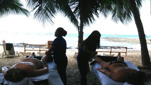 Un salon de massage situé en bord de mer, avec deux tables de massage installées sous des palmiers, offrant une vue sur l’océan.