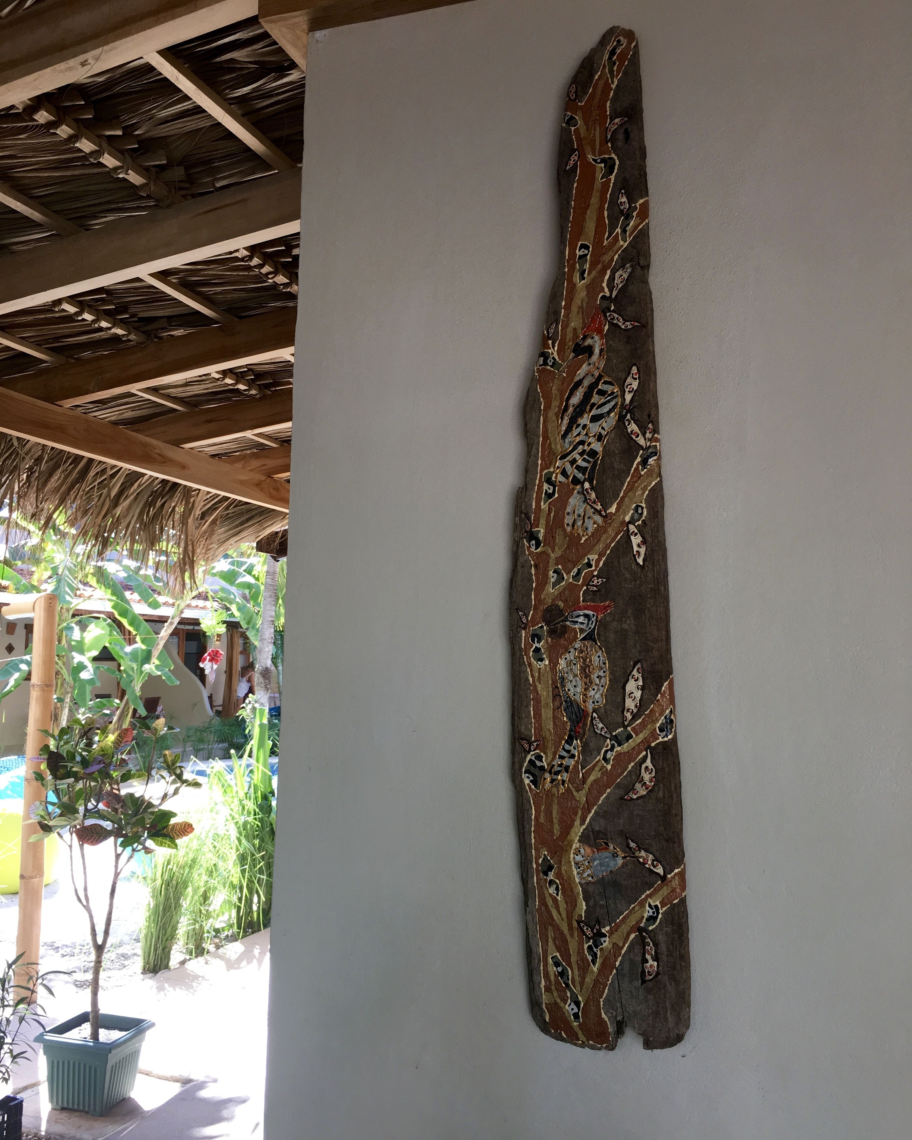 A long, vertically oriented wooden sculpture with intricate carvings of branches and leaves, mounted on a plain white wall.