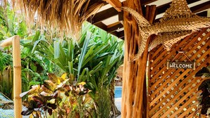 L’entrée d’un complexe hôtelier tropical, avec un toit de chaume, une végétation luxuriante et une pancarte indiquant « BIENVENUE ».
