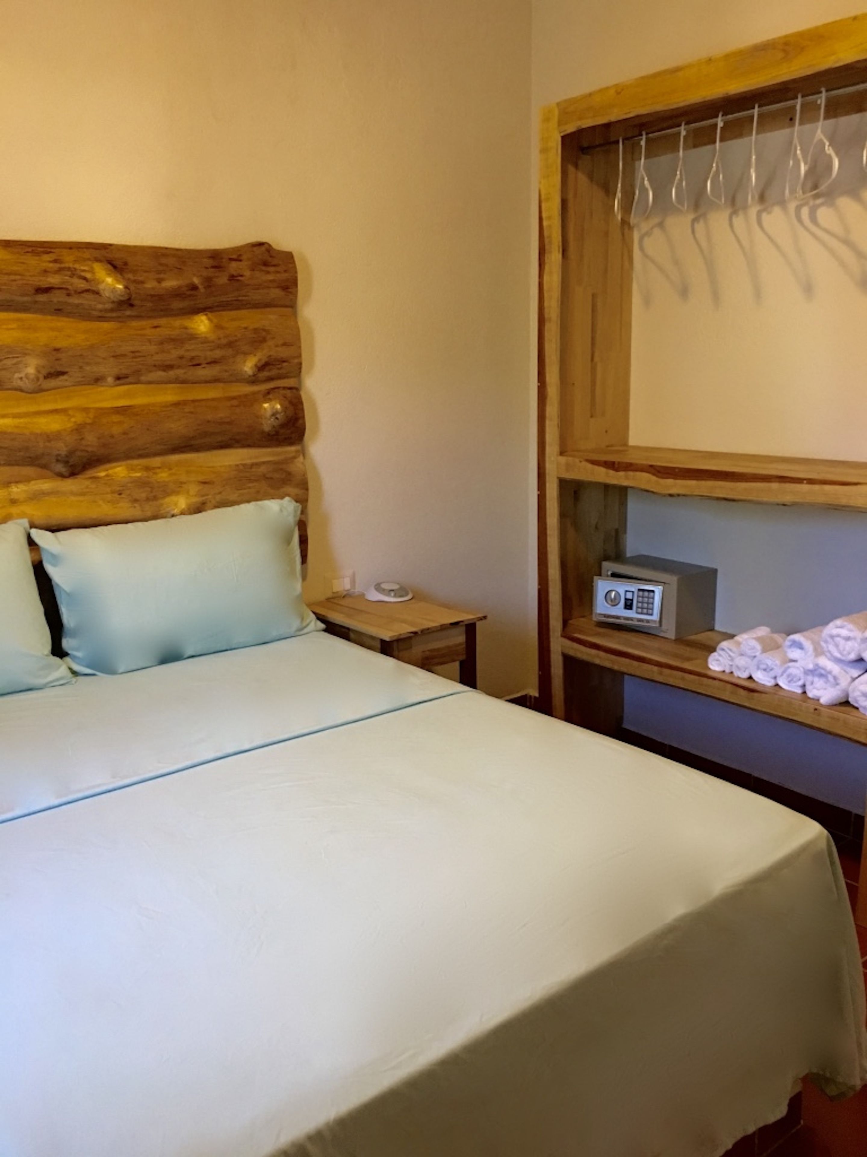 Uma cama com cabeceira de madeira, mesa de cabeceira com rádio e prateleira com toalhas.