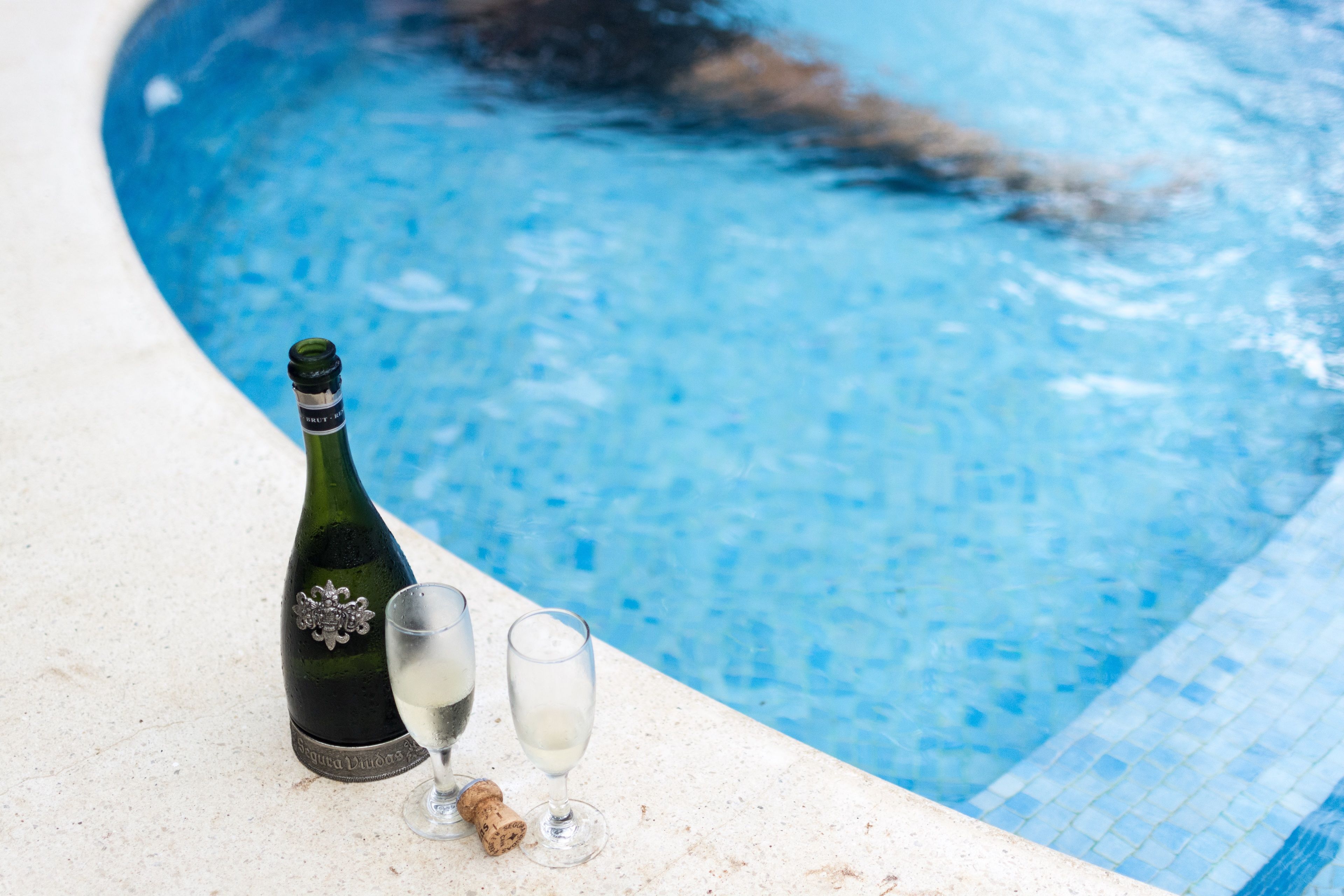 A poolside setting with a bottle of champagne and two glasses, one with a cork.