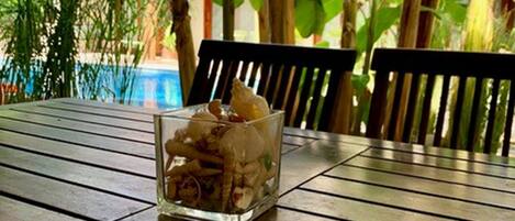 A glass container with seashells on a wooden table outdoors, surrounded by lush greenery and a building in the background.