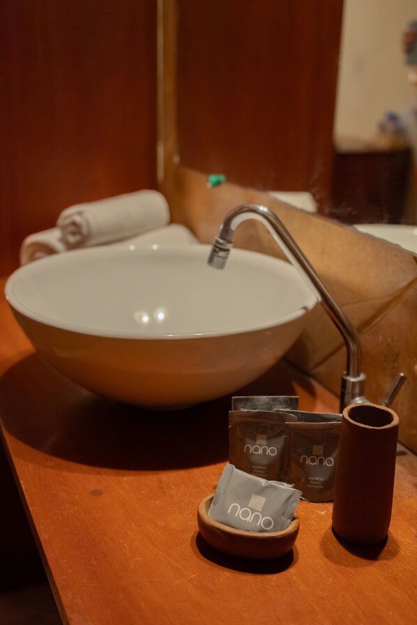 Un lavabo de baño con un recipiente grande, un grifo y una toalla enrollada a un lado. Hay dos pequeños recipientes con el nombre de marca