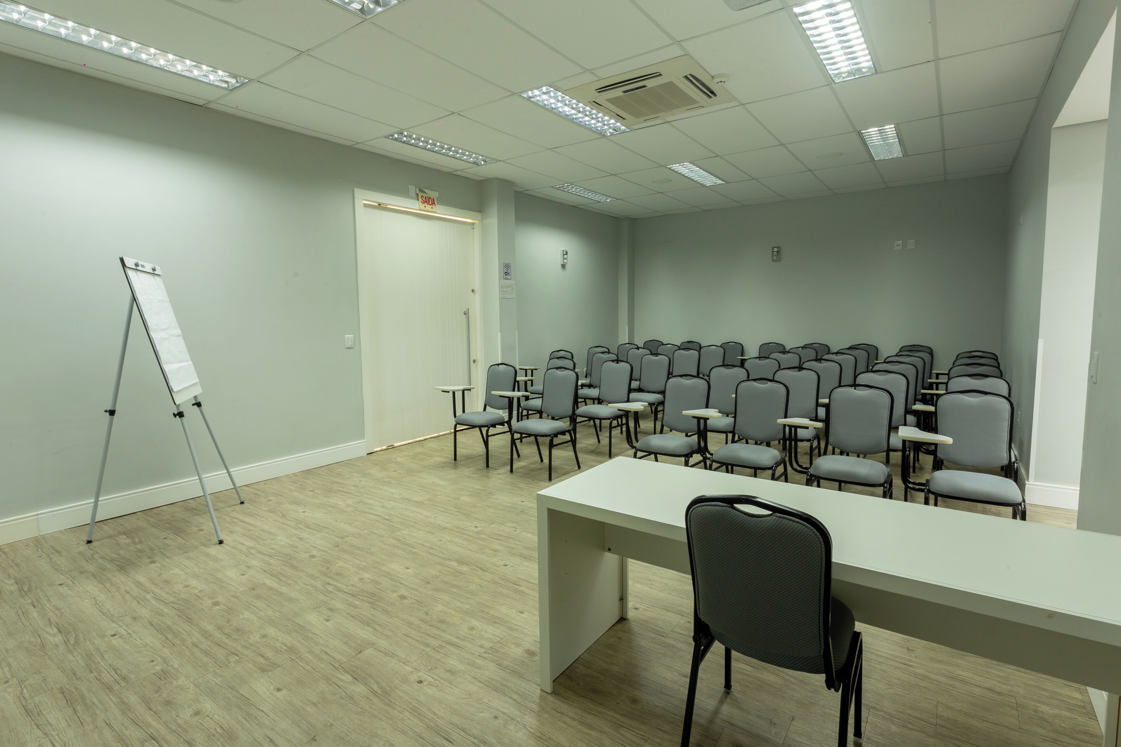 Una sala de conferencias con una mesa larga, varias sillas, un soporte para pizarra blanca y una puerta con la inscripción 'EXIT'.
