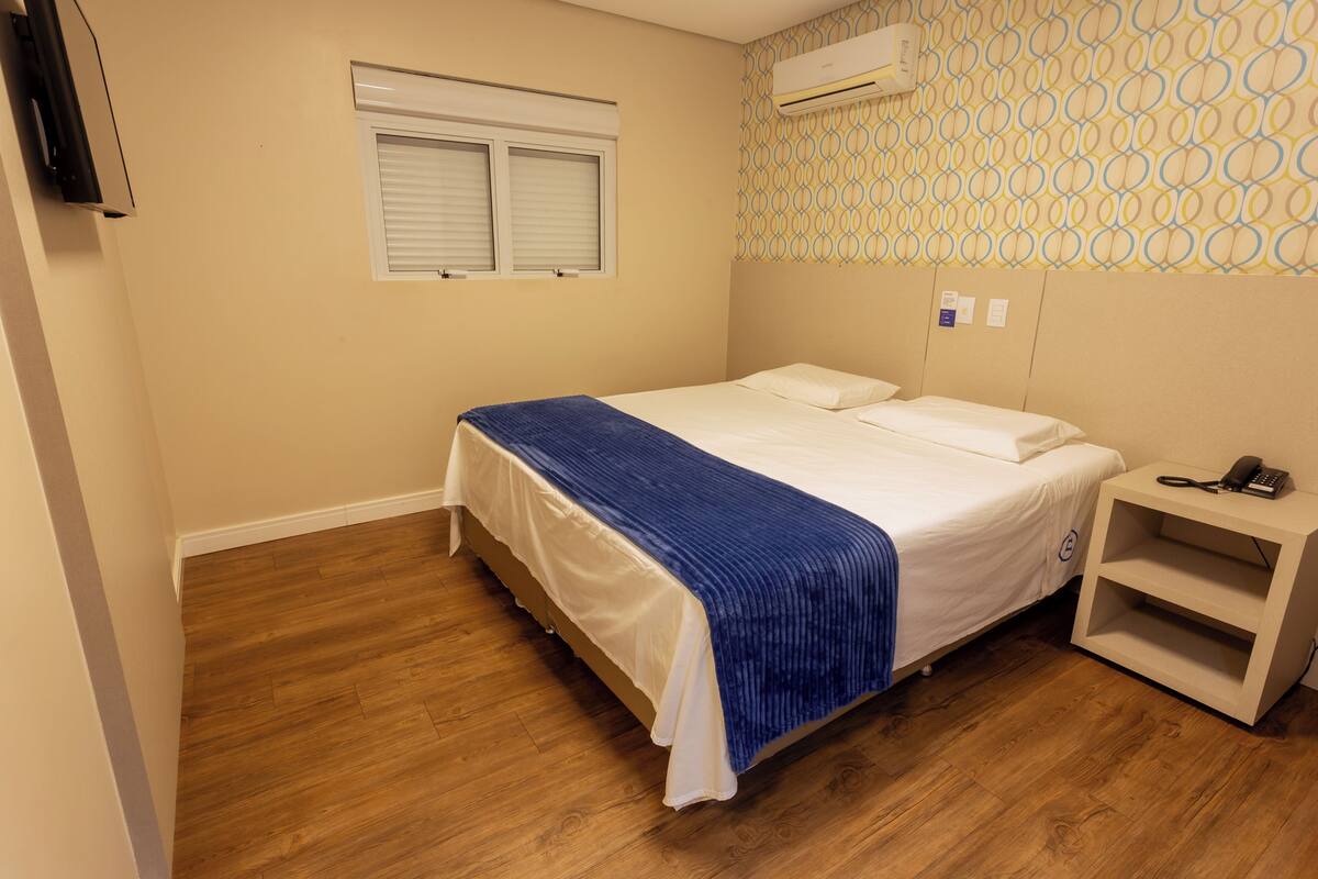 Ein Schlafzimmer mit Bett, Nachttisch, Wand-Klimaanlage, einem Fenster mit Jalousien und einer Wand mit Muster.