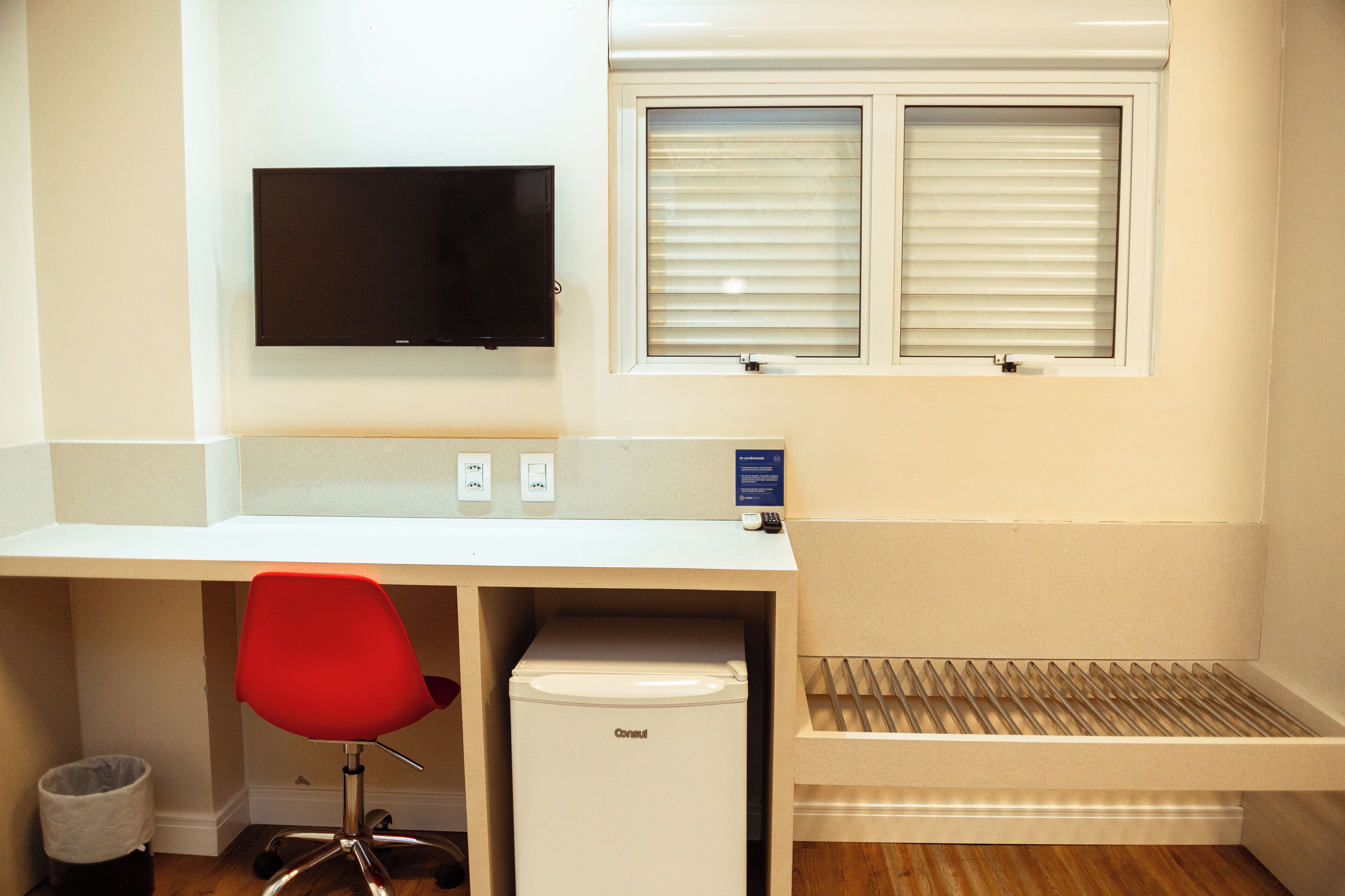 Una habitación con un escritorio blanco, una silla roja, un refrigerador blanco pequeño, un televisor montado y una ventana con persianas.