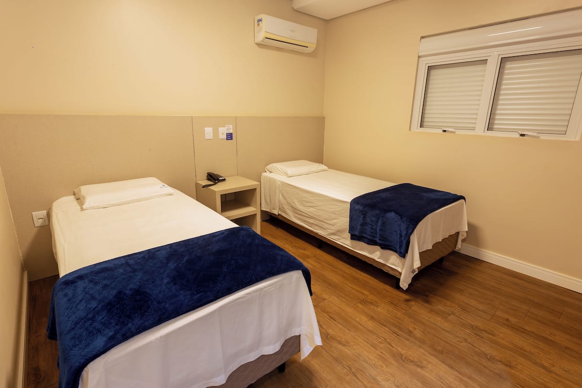 Ein Hotelzimmer mit zwei Einzelbetten, einem Nachttisch mit Telefon, einer Klimaanlage und einem Fenster mit Jalousien.