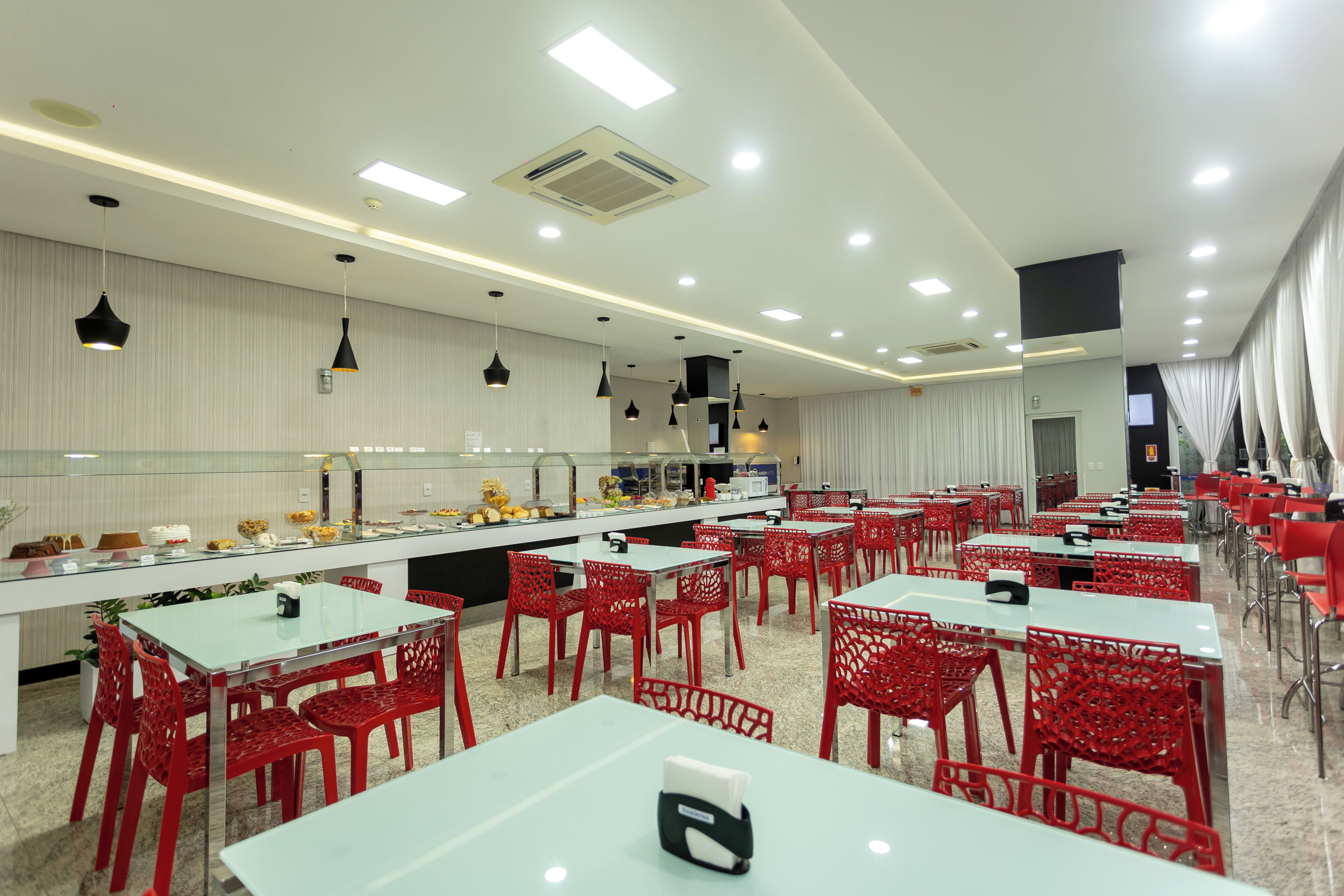 Un espace repas moderne avec des tables blanches et des chaises rouges, un buffet proposant divers plats, et un éclairage encastré au plafond.