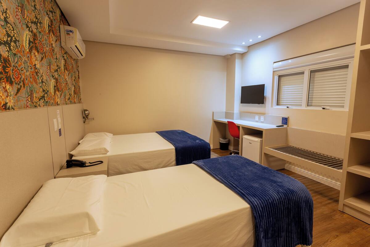 Ein Hotelzimmer mit zwei Betten, einem Schreibtisch, einem Fernseher und einem Fenster mit Jalousien.