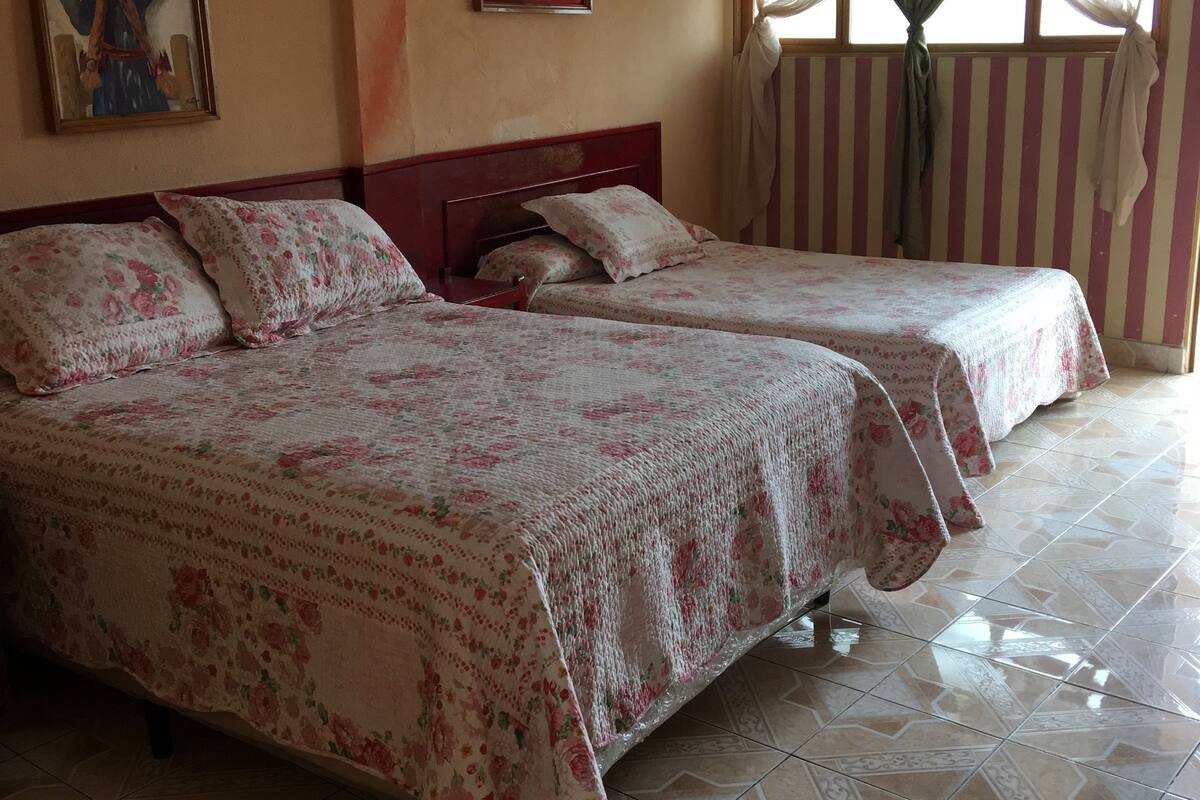 Une chambre d’hôtel avec deux lits, des couvre-lits à motifs floraux, une grande fenêtre avec des rideaux et un sol carrelé.