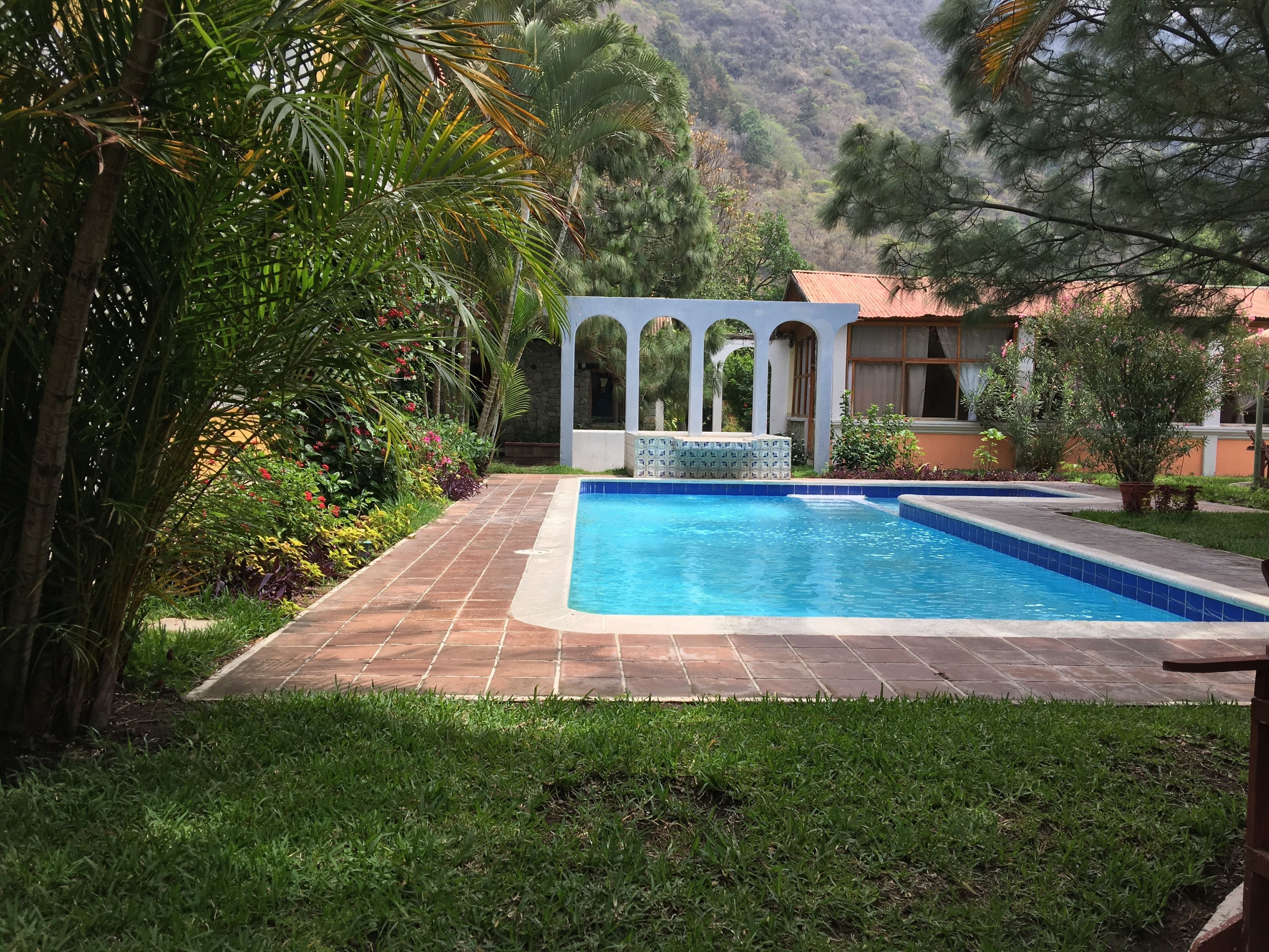 A swimming pool with a tiled deck, surrounded by lush greenery and a stone building with arches.
