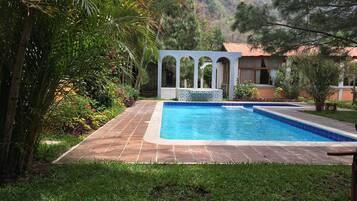 A swimming pool with a tiled deck, surrounded by lush greenery and a stone building with arches.