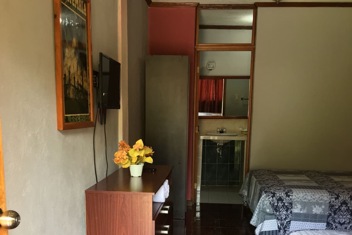 Une chambre d’hôtel avec un bureau en bois, un lit avec une literie à motifs, une salle de bain avec un lavabo et une affiche accrochée au mur.
