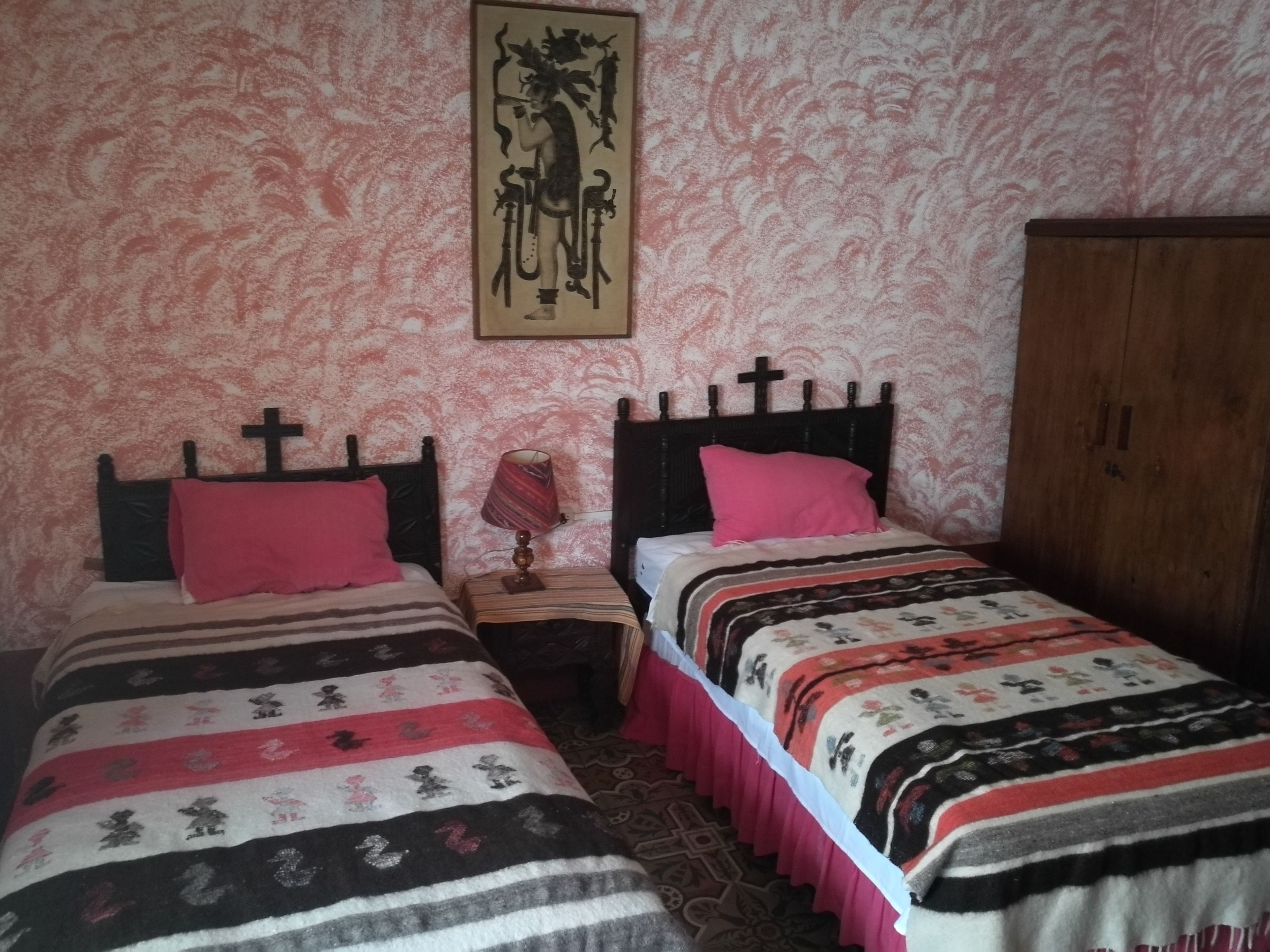 A bedroom with two beds, each with a cross headboard, a patterned wall, and a wooden wardrobe.
