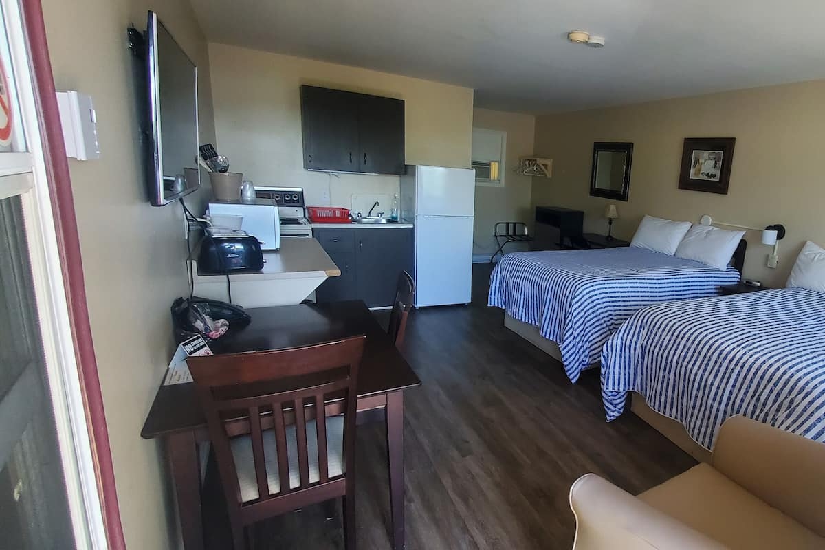 A compact hotel room with two beds, a kitchenette, a desk with a chair, and a small seating area.
