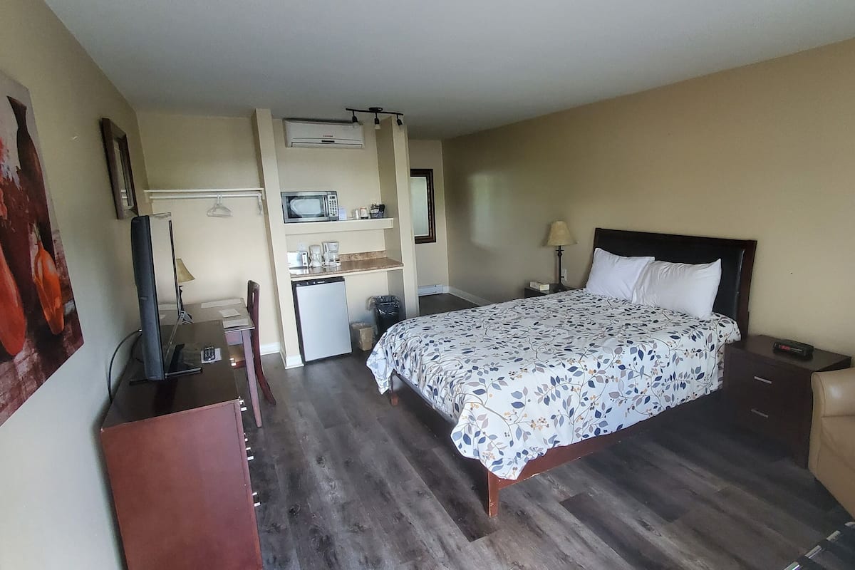 A hotel room with a bed, a desk with a computer, a chair, a small kitchenette, and a large window.