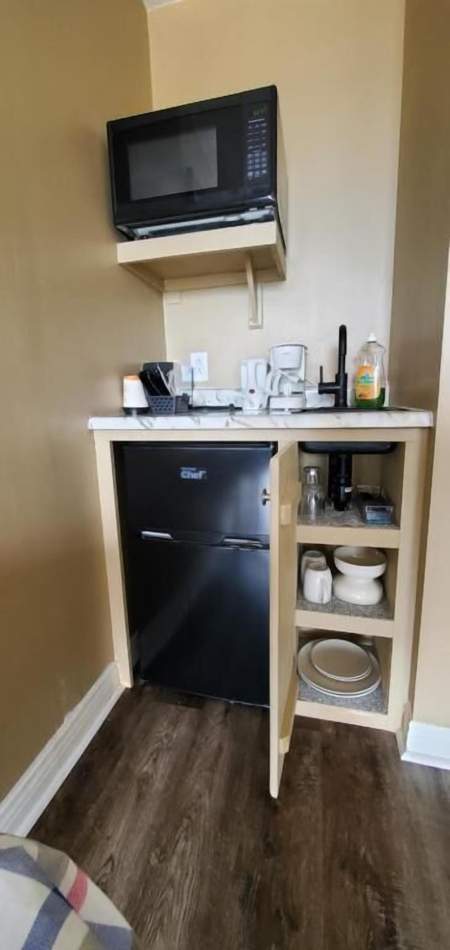 Una kitchenette compacta equipada con microondas, refrigerador, cafetera y una alacena con vajilla.