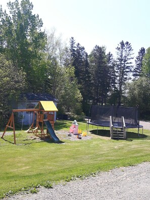 Une aire de jeux avec des balançoires, un toboggan et un trampoline, située dans un espace verdoyant avec des arbres en arrière-plan.