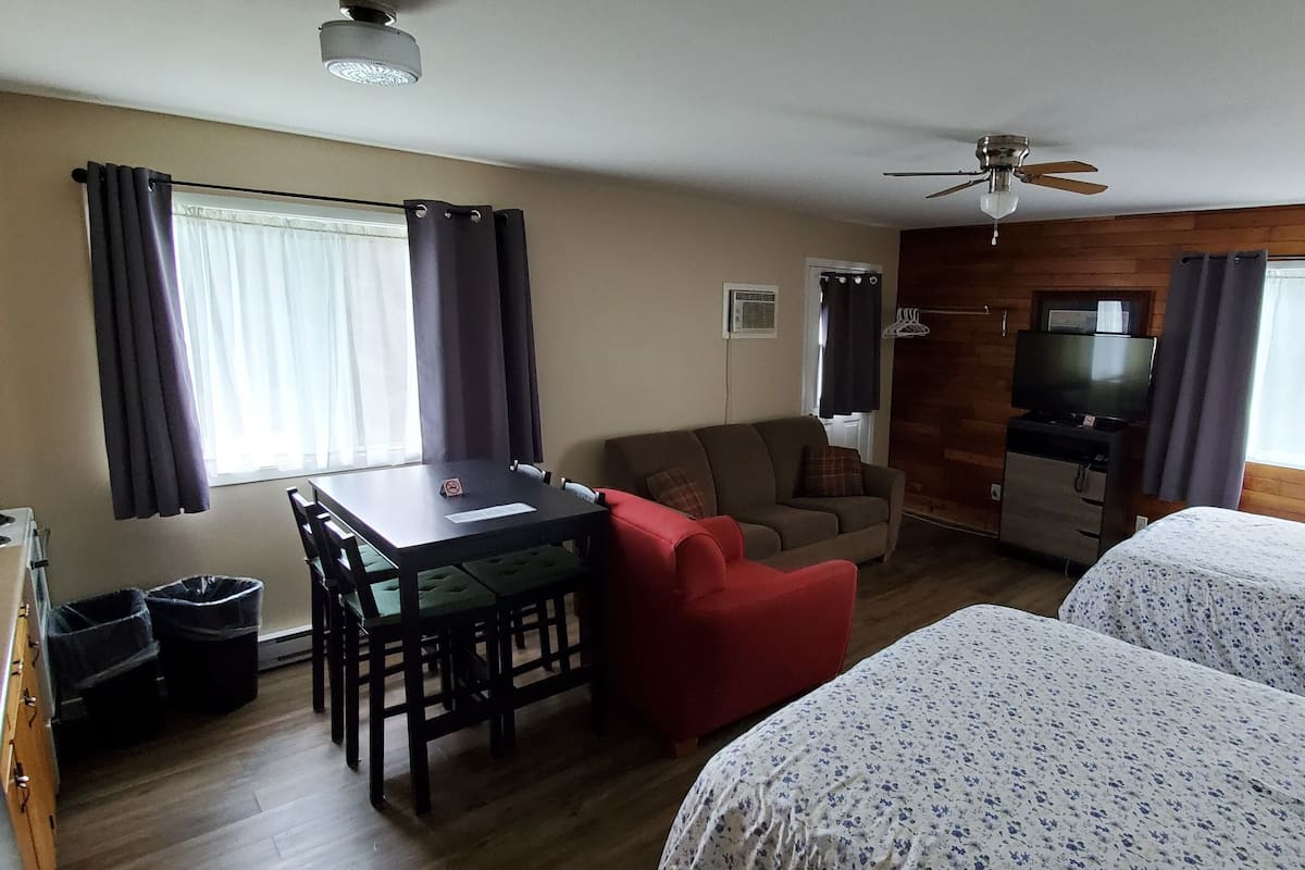 A hotel room with a wooden wall, a TV, a desk with chairs, two beds, and a ceiling fan.