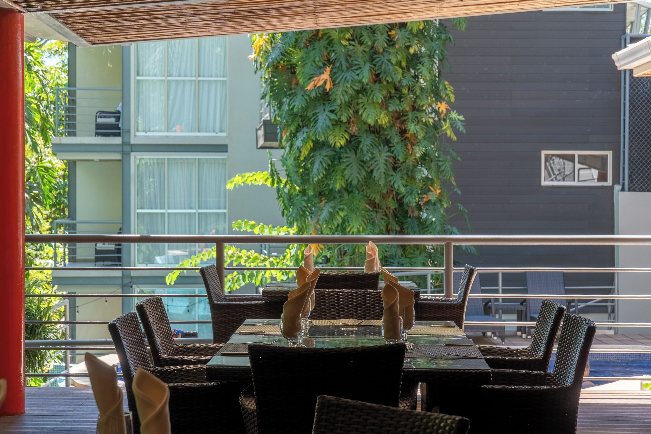 An outdoor dining area with wicker furniture and glass tables, overlooking a building with large windows and a green wall.