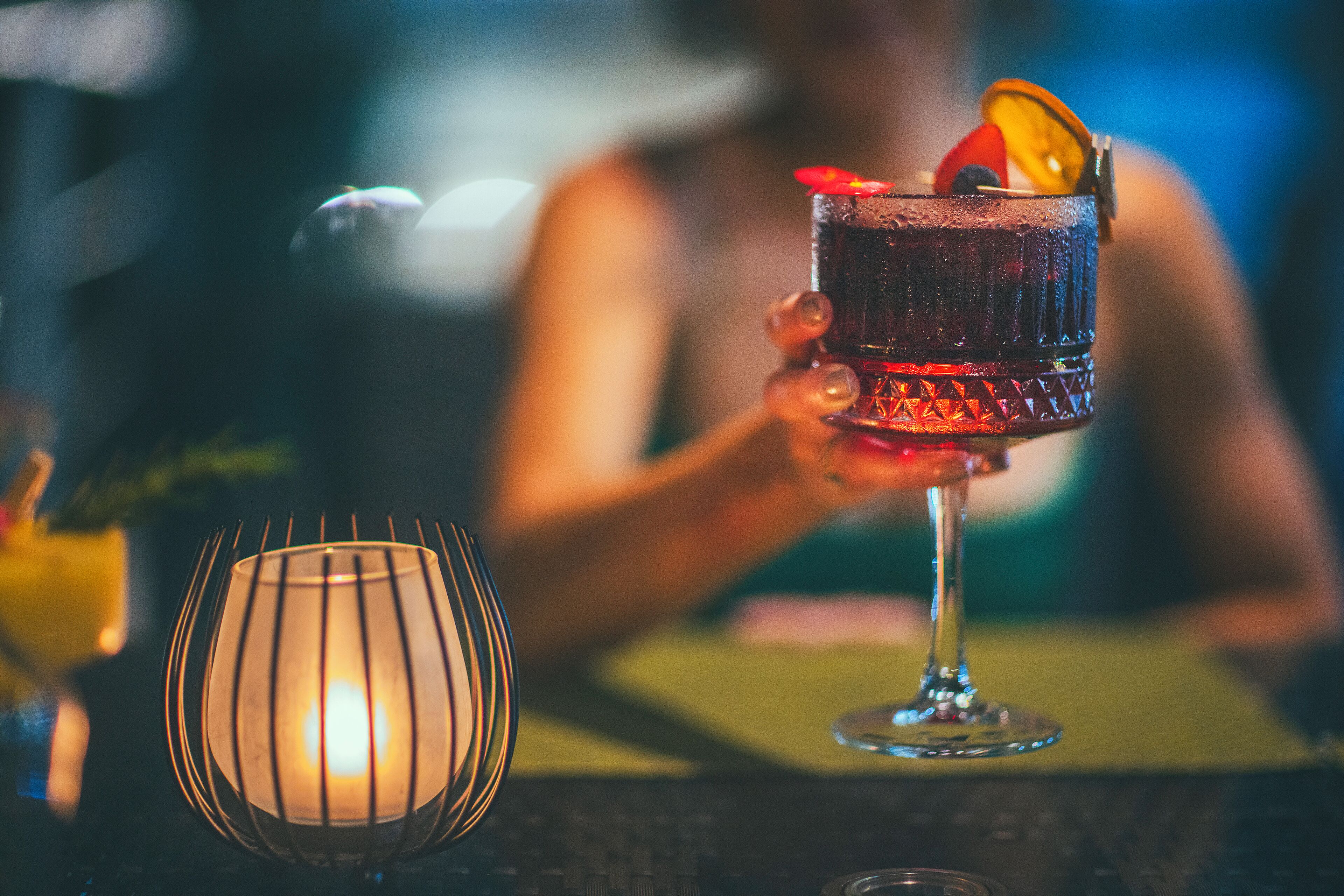 Un verre à cocktail, garni d’une tranche d’orange et d’un fruit rouge, tenu par une personne.