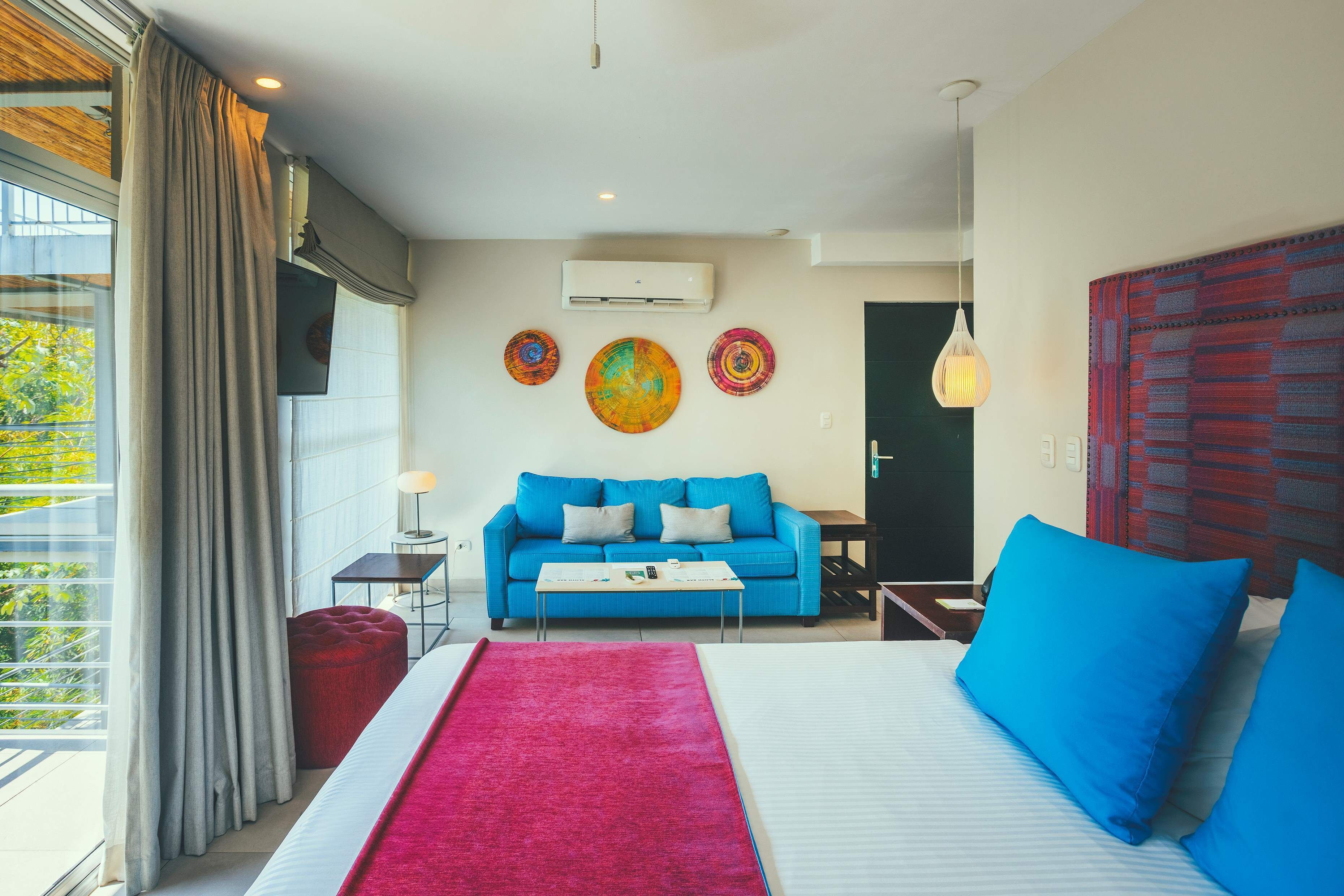 Ein modernes Hotelzimmer mit einem großen Bett, einem blauen Sofa und einem roten Hocker. Ein Fenster mit Vorhängen, eine wandmontierte Klimaanlage und ein farbenfrohes Kunstwerk über dem Sofa sind ebenfalls vorhanden.