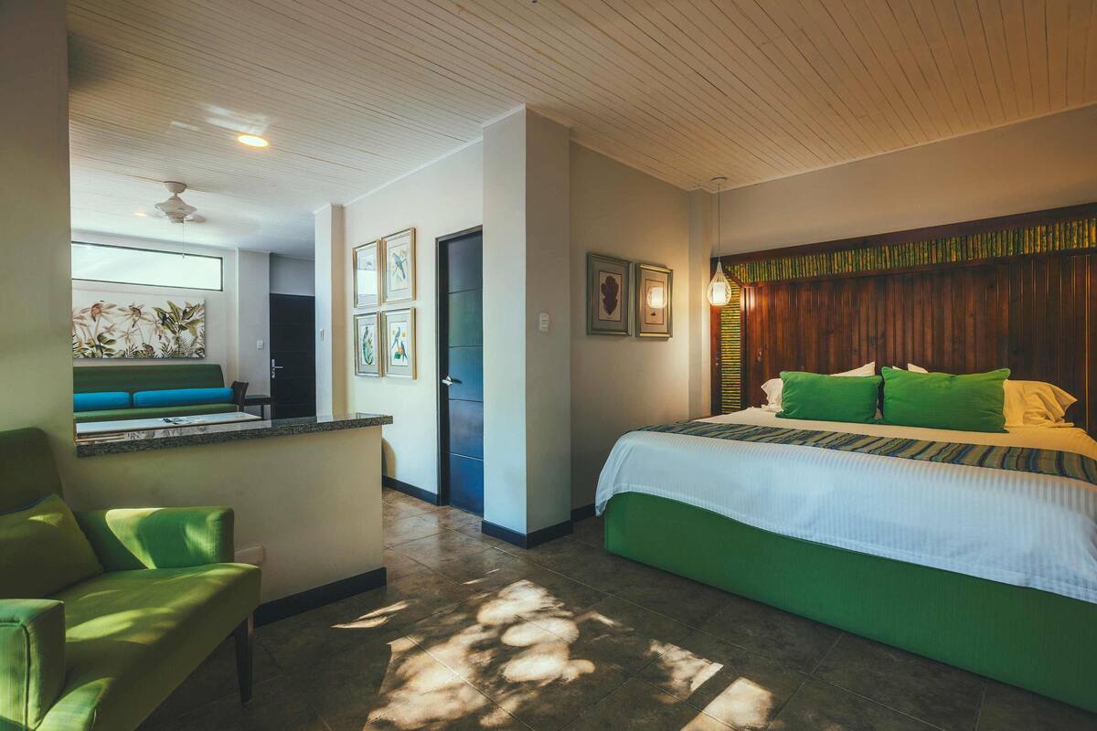 Habitación de hotel con una cama, un sillón verde, un escritorio pequeño y una ventana con vistas al exterior.
