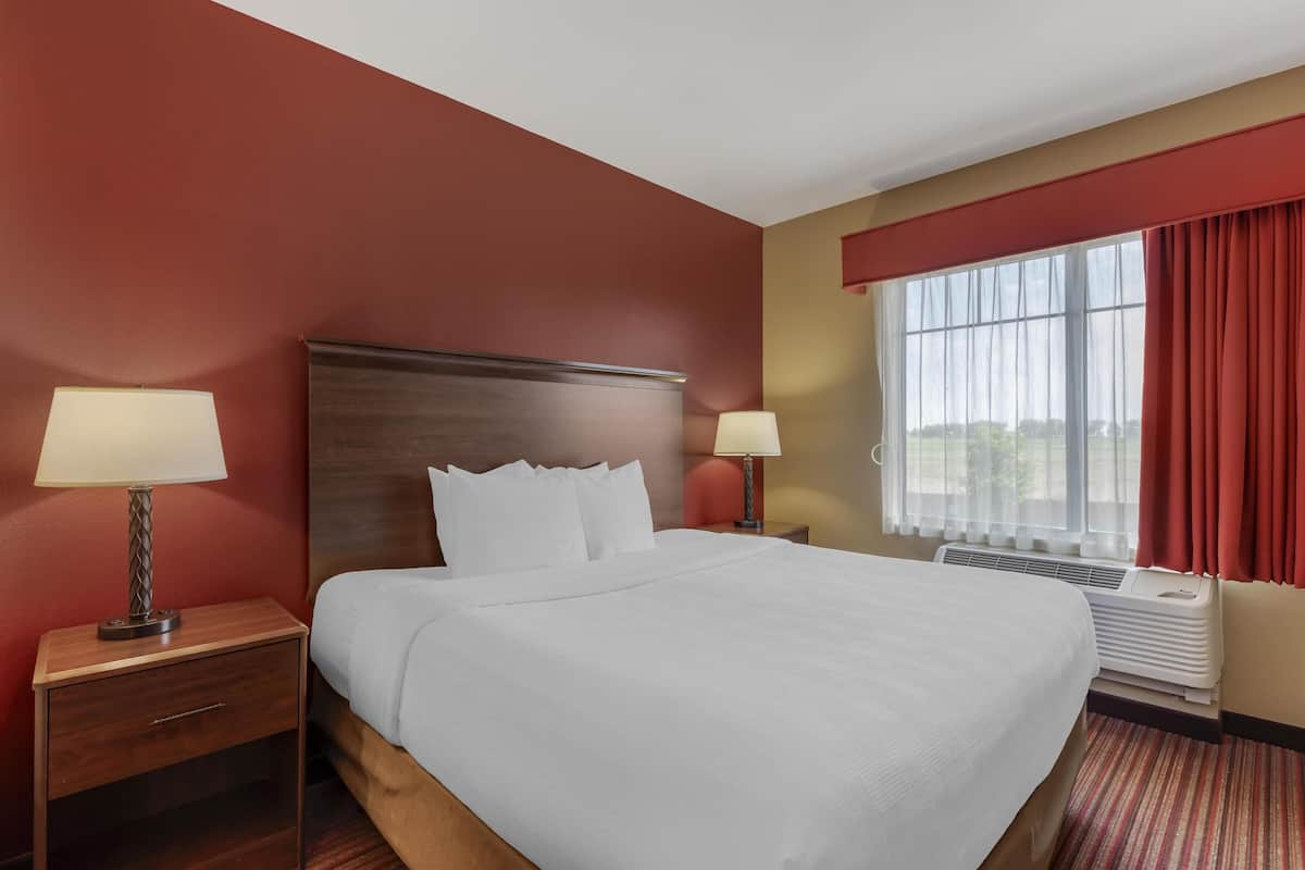 A hotel room with a large bed, two bedside tables with lamps, a window with red curtains, and a view of the outdoors.