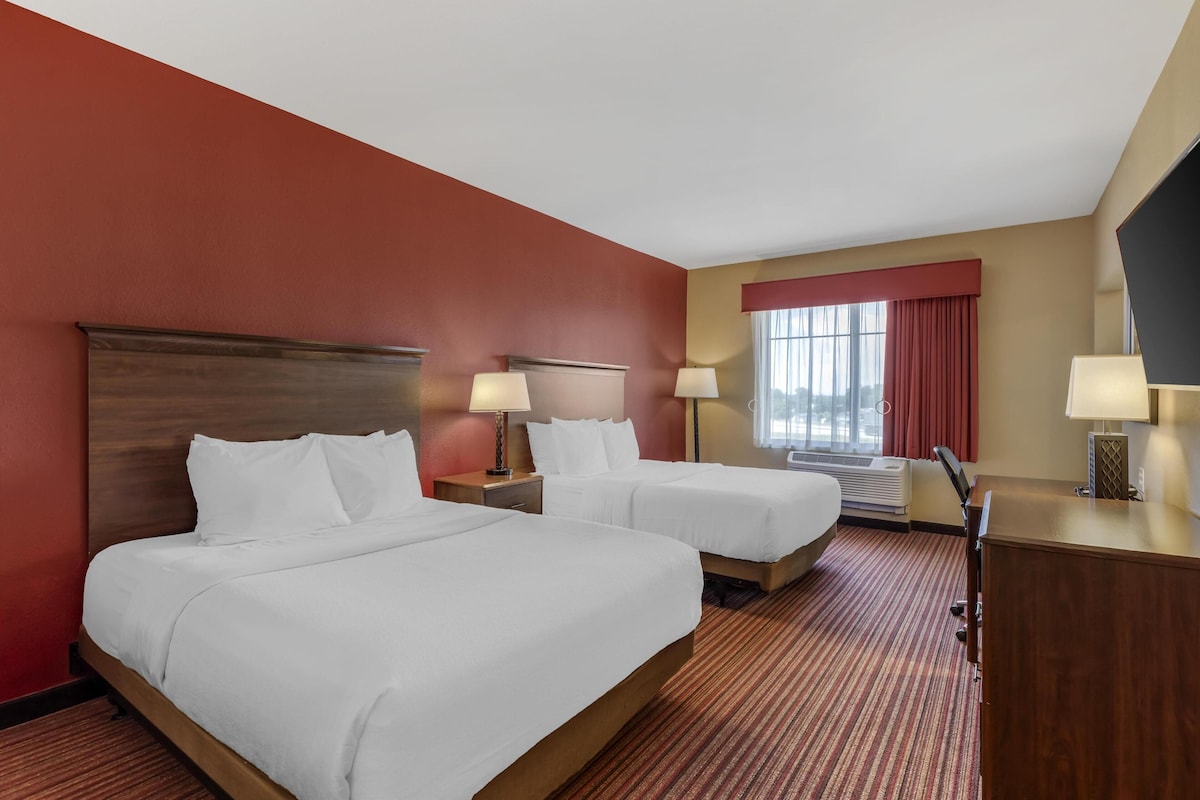 A hotel room with two beds, a wooden headboard, a red wall, a window with red curtains, and a desk with a lamp.