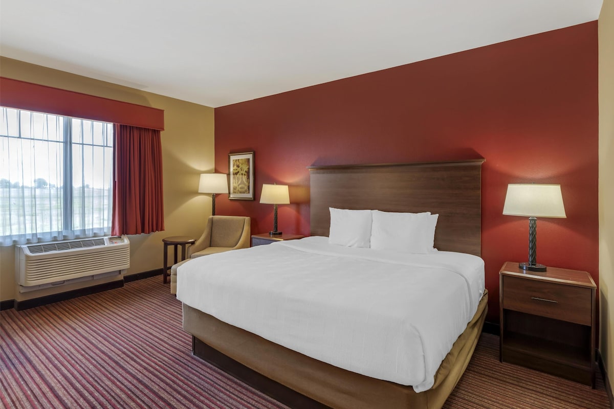 A hotel room with a large bed, two bedside tables with lamps, a chair, a window with curtains, and a wall-mounted air conditioner.