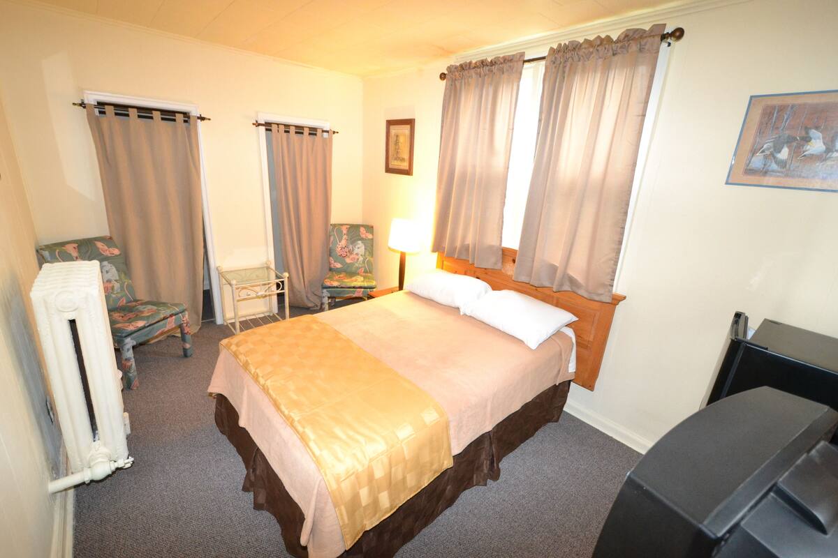 A hotel room with a bed, a chair, a desk, a lamp, and a radiator.