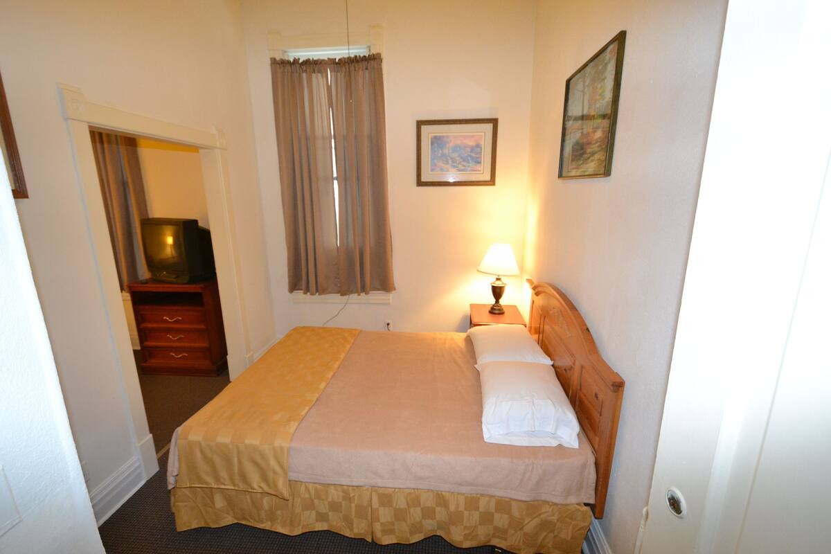 A hotel room with a bed, a nightstand with a lamp, a wooden dresser, a television, and a window with curtains.