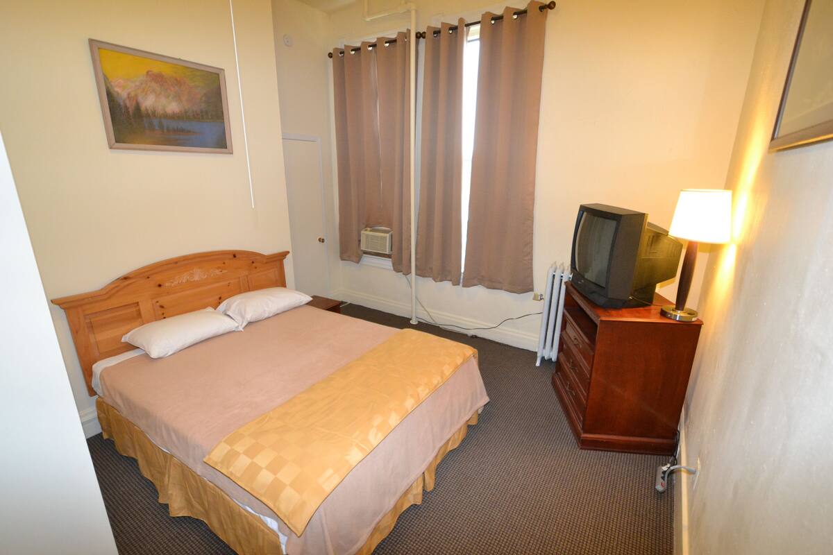 A hotel room with a single bed, a television on a wooden dresser, a painting on the wall, and a window with curtains.