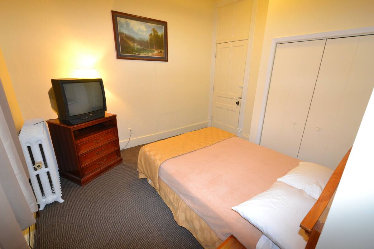 A small, neatly arranged bedroom with a single bed, a wooden nightstand, a television on a stand, and a radiator.