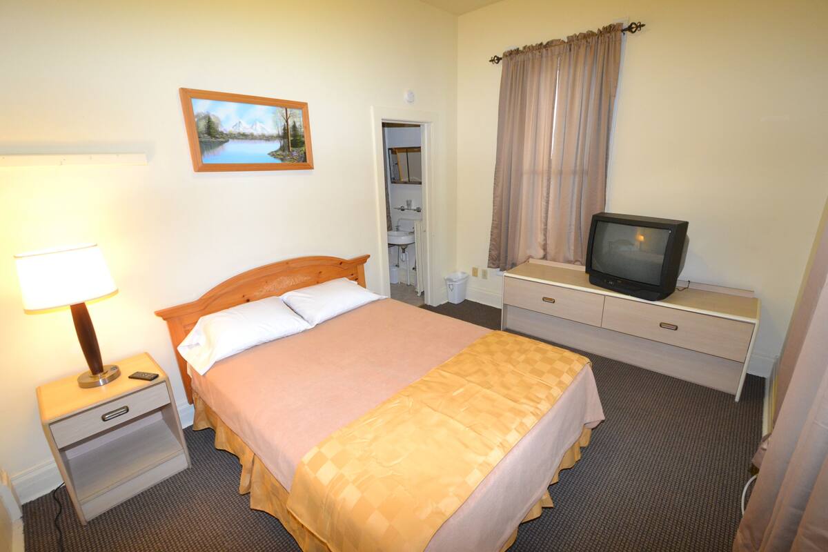 A neatly arranged hotel room with a bed, bedside table with a lamp, a television on a dresser, and a painting on the wall.