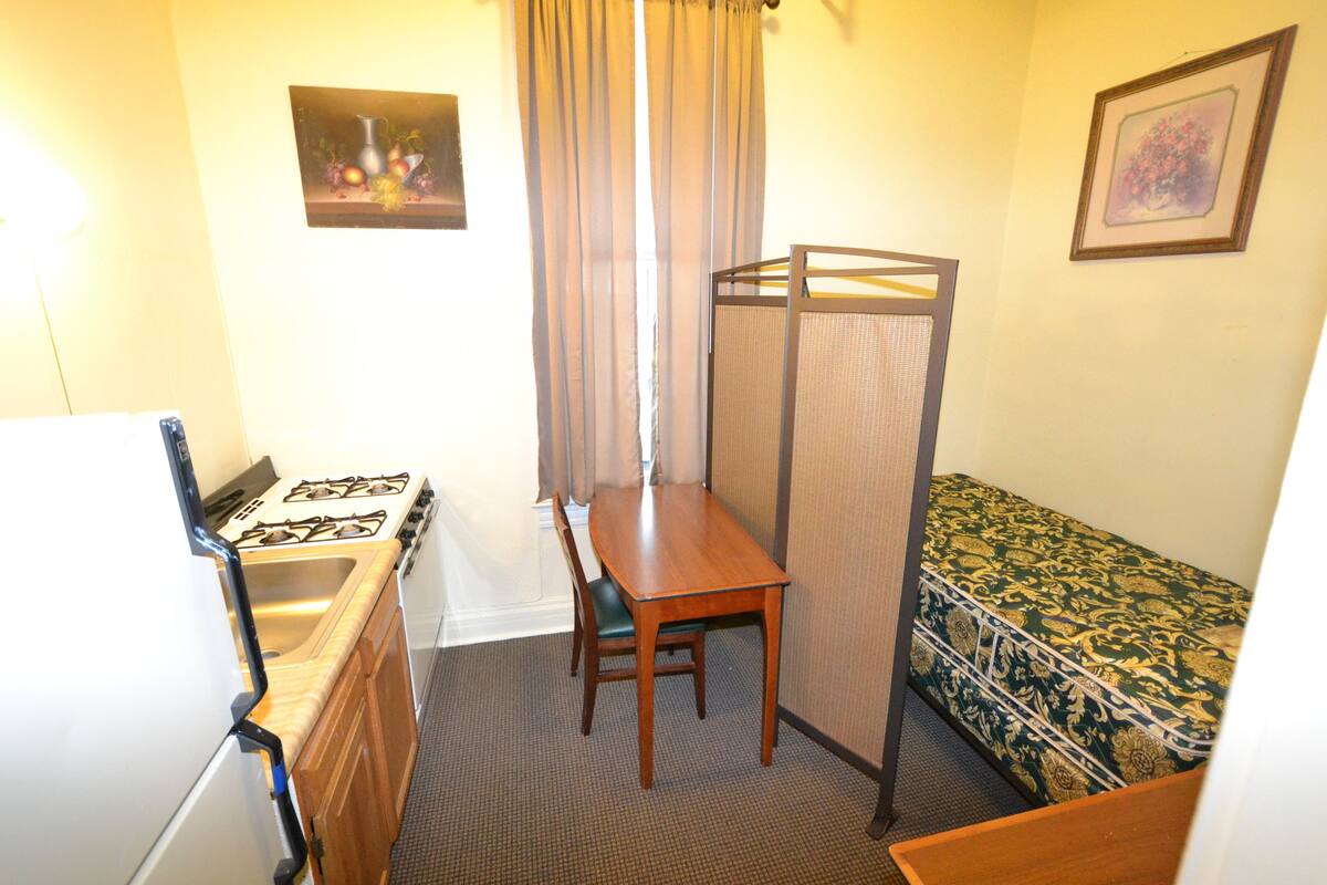 A compact room with a kitchenette, a single bed, a dining table, and a chair.