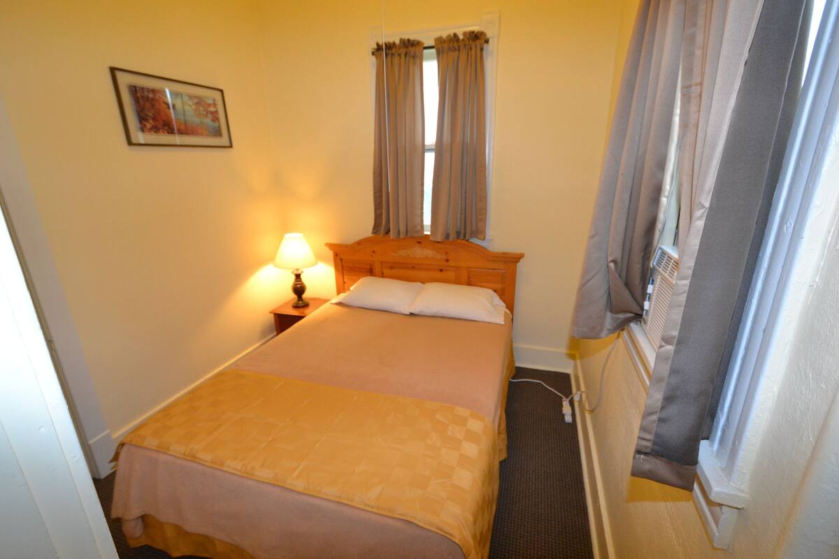 A hotel room with a bed, a nightstand with a lamp, a window with curtains, and a painting on the wall.